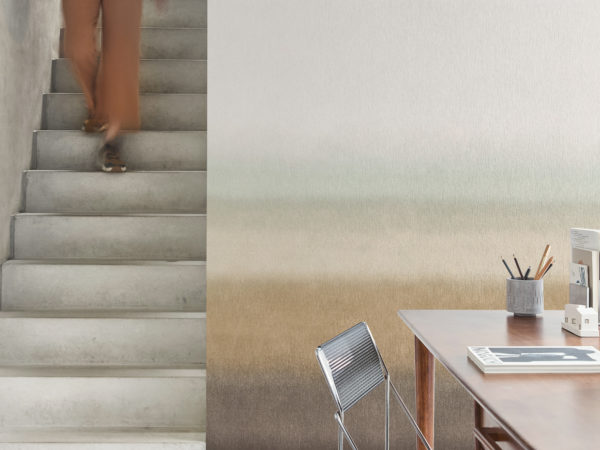 A person wearing a blue top and brown pants is descending a minimalist concrete staircase on the left side of the image. The background wall features Omexco’s printed non-woven “Edition” collection, a timeless and soft gradient design transitioning from from beige, light green and light brown to dark brown. On the right, there is a modern wooden desk with a few items, including a magazine, a cup with pencils, and a small decorative object. A sleek metal chair is positioned in front of the desk. The floor is light-colored, complementing the overall modern and minimalist aesthetic of the space.
