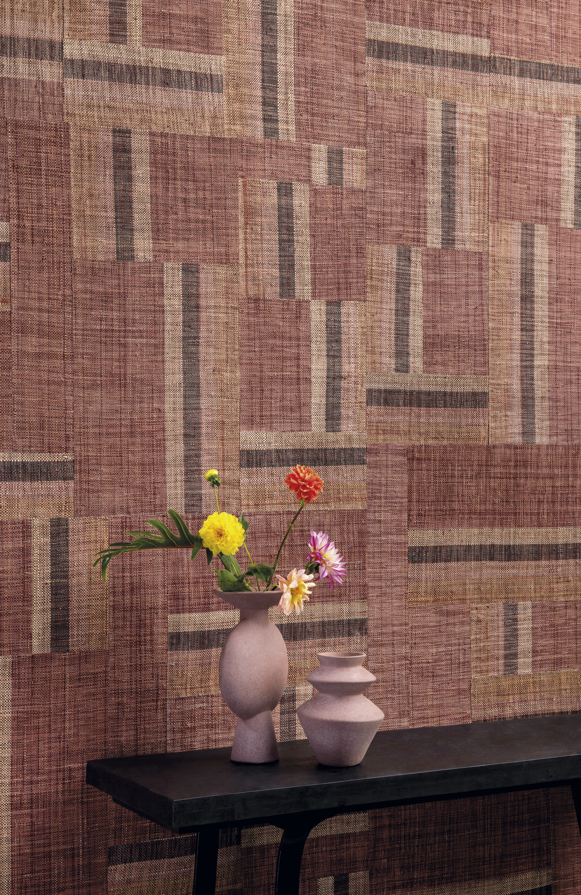 Authentische Handwerkskunst verkörpert die kühne Kollektion Atelier, die von den kreativen Köpfen von Omexco entworfen wurde. Dieses Bild zeigt ein beeindruckendes rot-braunes Patchwork aus handgewebtem Raffia. Die perfekte Kulisse für einen hölzernen Beistelltisch mit zwei rosafarbenen Keramikvasen und einigen frischen Feldblumen.