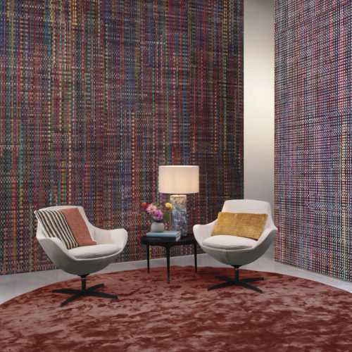 Vom Webstuhl der Handweber in Ihr Wohnzimmer. An den Wänden dieses beeindruckenden Wohnzimmers sehen wir die Wandbekleidung "Extravaganza" aus der Atelier-Kollektion von Omexco. Bunte Bänder aus recycelter Sari-Seide sind mit Abaca handgewebt. Die gemütliche Sitzecke ist außerdem mit einem runden bordeauxfarbenen Teppich, einem Beistelltisch mit Lampe, zwei weißen Loungesesseln mit bunten Kissen und einem kleinen Strauß frischer Schnittblumen dekoriert.