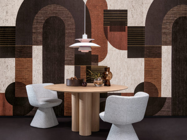 Beispiel für ein Esszimmer mit einer kühnen grafischen Wandbekleidung, die ein Muster aus geometrischen Formen in grauen, schwarzen, orangefarbenen und weißen Farbtönen zeigt. Dieses Panoramamuster ist digital auf Kork gedruckt und gehört zur Omexco-Wandbelagskollektion "Casca". In der Mitte des Raumes steht ein runder Esstisch aus Holz, über dem eine weiße Hängelampe hängt. Dekorative Elemente wie kleine Glasvasen und eine mit Walnüssen gefüllte Schale sind auf der Tischplatte platziert. Zwei weiche graue Esszimmerstühle sind um den Tisch herum angeordnet. Die Gesamtästhetik ist modern mit einem Hauch von Wärme und Textur.