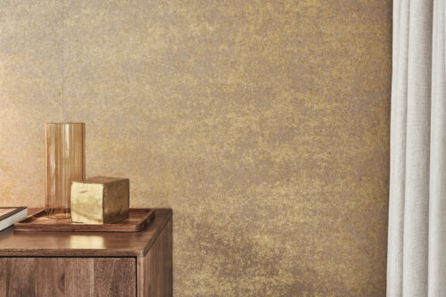 Eine Zimmerecke mit einer Patina-Effekt-Wandbekleidung aus der gedruckten Vlieskollektion "Edition" von Omexco. Die Wand hat eine strukturierte gold- und braunfarbene Oberfläche. Im Vordergrund steht ein Holzschrank mit einem dekorativen Tablett, auf dem eine hohe, bernsteinfarbene Glasvase und ein goldfarbener Würfel stehen. Rechts ist ein Blick auf hellfarbene Vorhänge zu sehen.
