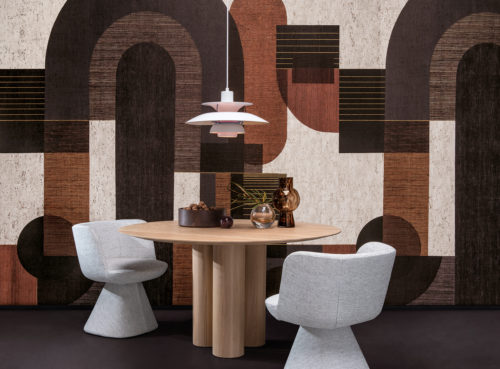 Beispiel für ein Esszimmer mit einer kühnen grafischen Wandbekleidung, die ein Muster aus geometrischen Formen in grauen, schwarzen, orangefarbenen und weißen Farbtönen zeigt. Dieses Panoramamuster ist digital auf Kork gedruckt und gehört zur Omexco-Wandbelagskollektion "Casca". In der Mitte des Raumes steht ein runder Esstisch aus Holz, über dem eine weiße Hängelampe hängt. Dekorative Elemente wie kleine Glasvasen und eine mit Walnüssen gefüllte Schale sind auf der Tischplatte platziert. Zwei weiche graue Esszimmerstühle sind um den Tisch herum angeordnet. Die Gesamtästhetik ist modern mit einem Hauch von Wärme und Textur.