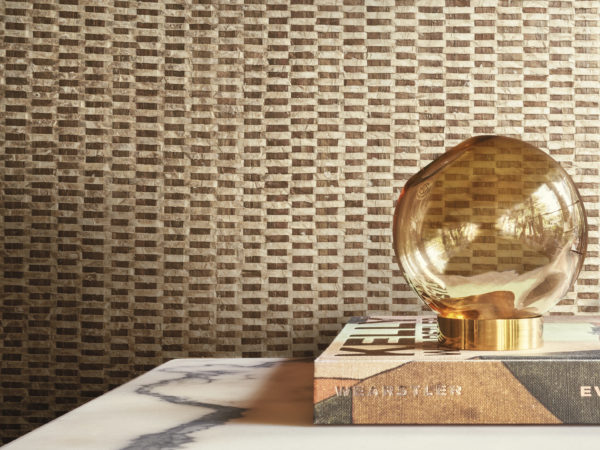 Une élégante lampe de table ronde en verre, joliment placée au-dessus d'un livre, sert de point focal à ce décor élégant. La toile de fond est constituée de luxueux revêtements muraux en capiz de la collection Archipelago d'Omexco, qui présentent un mélange captivant de tons dorés et bruns, créant ainsi une atmosphère sophistiquée et accueillante.