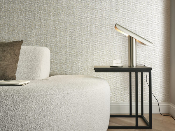 Gros plan sur un salon confortable, avec un canapé en tissu bouclé beige orné d'un coussin marron. Le parquet à chevrons complète les plinthes classiques, créant une esthétique intemporelle. Une table d'appoint noire se trouve à proximité, ornée d'une lampe de table dorée minimaliste. En toile de fond, les revêtements muraux en nacre capiz de la collection Archipelago d'Omexco présentent des nuances chatoyantes de beige, ajoutant une touche d'élégance et de sophistication à cet espace intime.