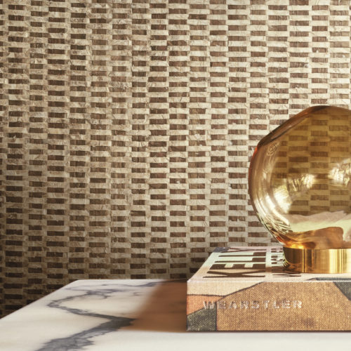 Une élégante lampe de table ronde en verre, joliment placée au-dessus d'un livre, sert de point focal à ce décor élégant. La toile de fond est constituée de luxueux revêtements muraux en capiz de la collection Archipelago d'Omexco, qui présentent un mélange captivant de tons dorés et bruns, créant ainsi une atmosphère sophistiquée et accueillante.